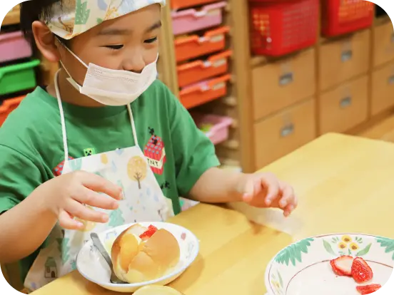 エプロンとマスク、バンダナを着用した男の子が、おやつ（または給食）のパンにイチゴをトッピングするクッキング活動を目の前にして、楽しそうに笑みを浮かべている様子