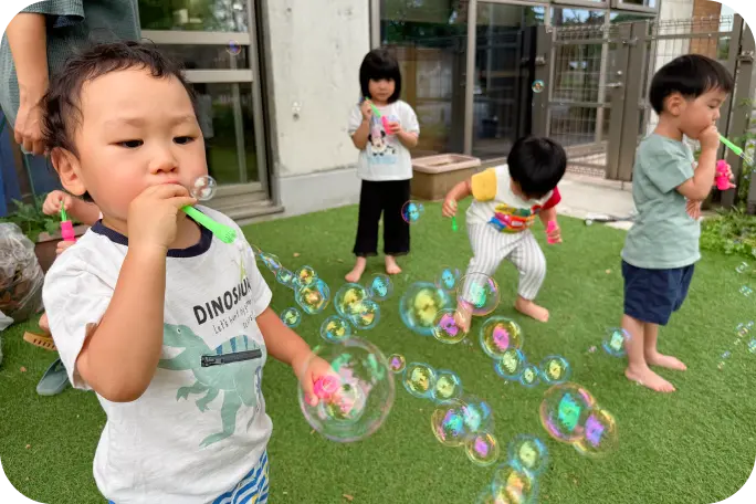 緑豊かな園庭の芝生の上で、手前の男の子が元気にシャボン玉を吹き、奥の園児たちも一緒にシャボン玉遊びを楽しんでいる様子