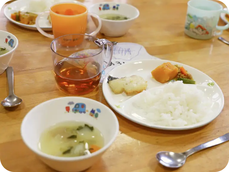 自園調理で手作りされた、温かいご飯、お魚とお野菜のおかず、具だくさんのスープ、お茶がセットされた園児向けの給食メニュー