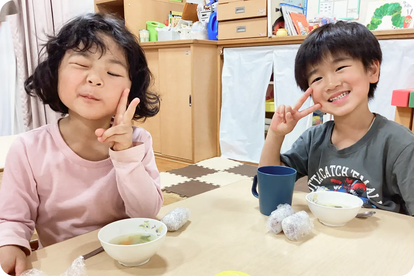 保育園の教室で、スープやおにぎりの食事を前にカメラに向かって笑顔でピースサインをする男の子と女の子