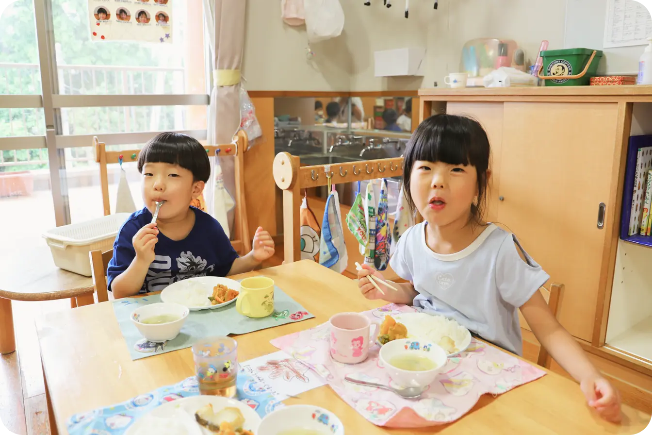 明るい保育室のテーブルを囲み、栄養バランスの行き届いた手作りの給食をおいしそうに笑顔で食べている男の子と女の子
