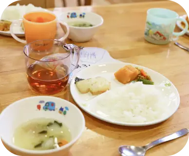 子ども用の車や動物のイラストが描かれた可愛い器に盛り付けられた、ご飯、焼き魚、かぼちゃの煮物、お味噌汁、お茶が並ぶつくしんぼ保育園の手作り給食