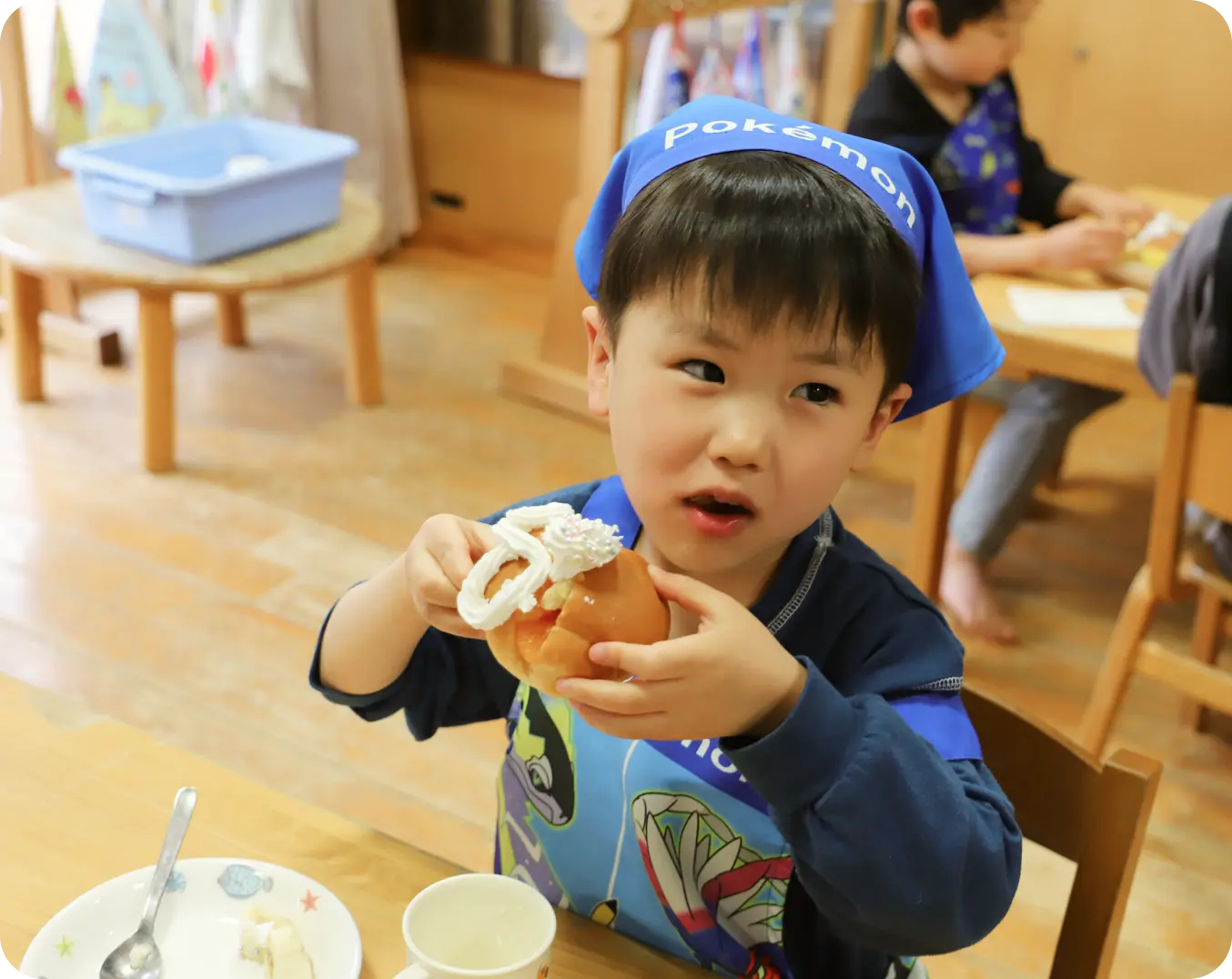 エプロンと青い三角巾を身につけ、生クリームが乗ったおやつのパンを両手で持って食べようとしている男の子