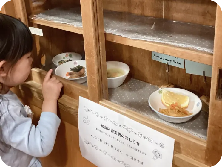 展示された当日の給食サンプルやお知らせの紙を、木製の棚のガラス越しに興味津々な様子で覗き込んでいるつくしんぼ保育園の女の子