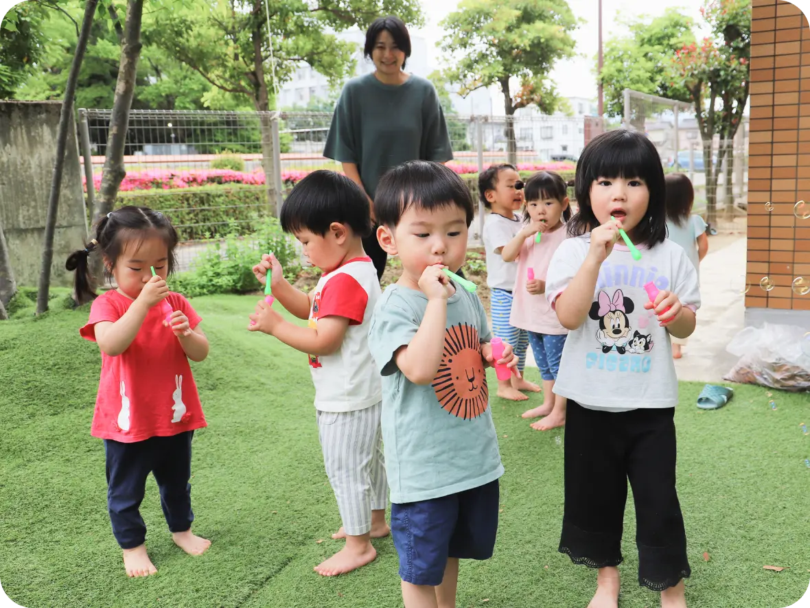 緑豊かな園庭の芝生の上で、保育士に見守られながら、思い思いにプラスチックのストローでシャボン玉を吹いて遊ぶ元気な園児たち