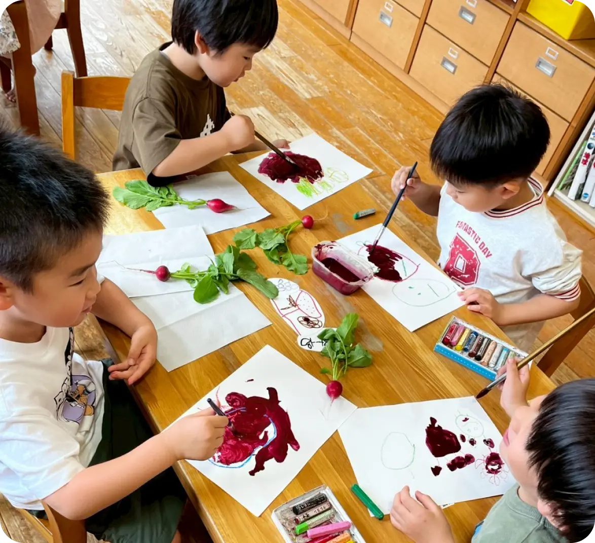 テーブルの上に置かれた本物の赤カブ（ラディッシュ）の葉や形をじっくり観察しながら、絵の具やクレヨンを使って自由に観察画を描く園児たち