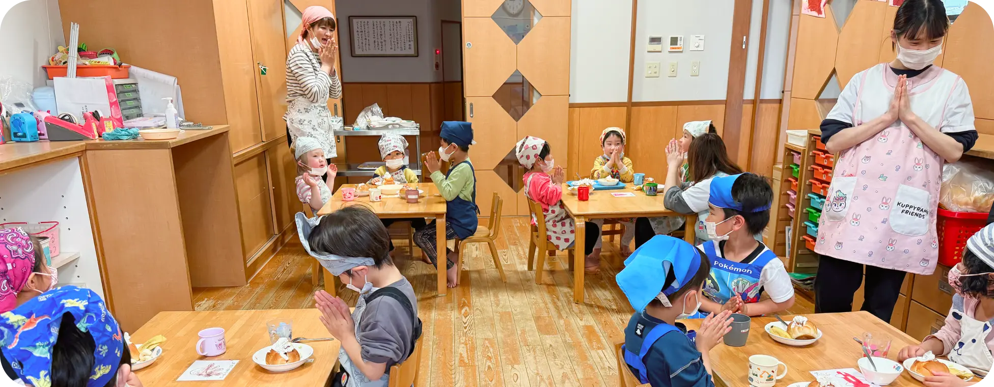 保育園の教室で、エプロンやバンダナを身につけた子どもたちと保育士が手を合わせて「いただきます」をするおやつの時間