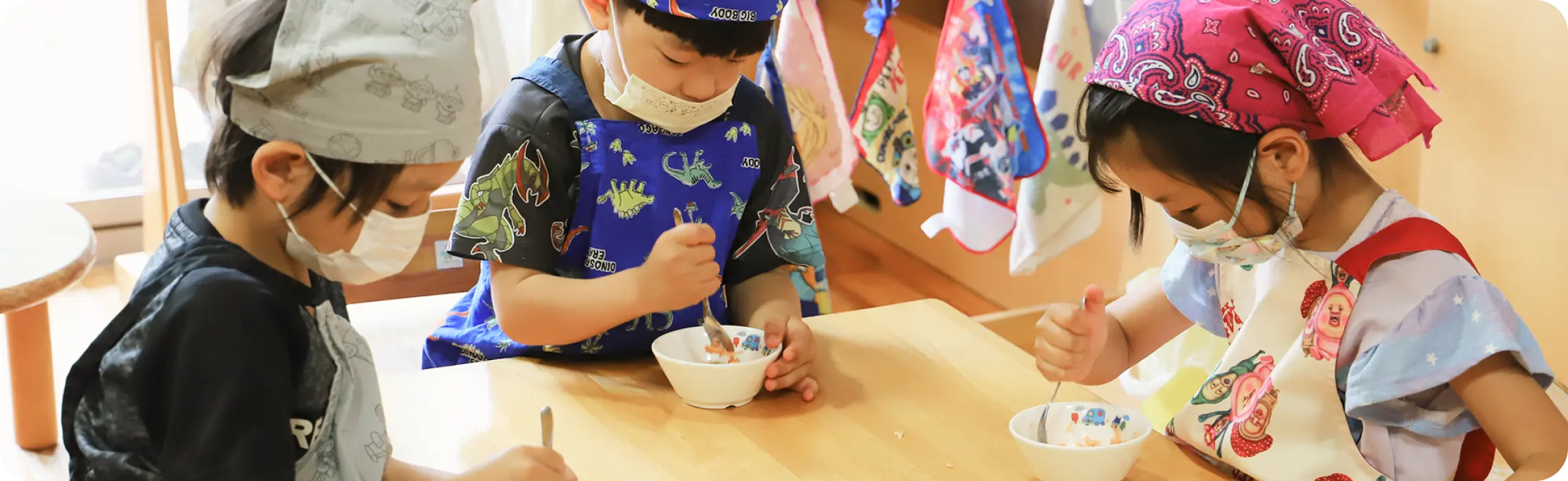 エプロンと三角巾、マスクを着用し、テーブルの上で自分の器に集中して丁寧におかずを盛り付けるつくしんぼ保育園の園児たち