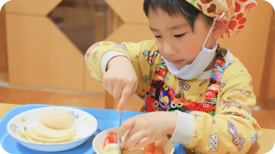 エプロン、三角巾、マスクを着用した男の子が、クッキング活動の中で自分の器にフォークを使って丁寧にフルーツを盛り付けているシーン