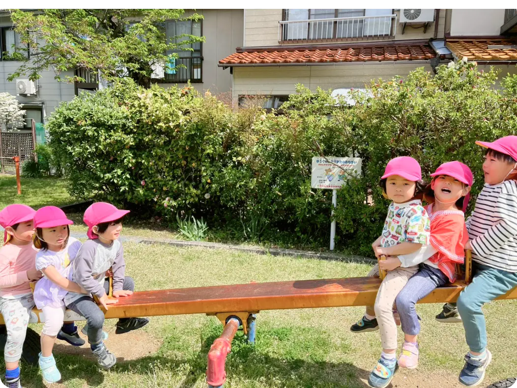 晴れた日に公園のシーソーに並んで座り、戸外遊びを楽しむ、ピンク色の帽子をかぶった子どもたちの様子