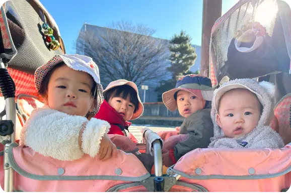晴れた日に帽子と防寒着を着用し、お散歩用の避難車に乗って外出する乳幼児たちの様子