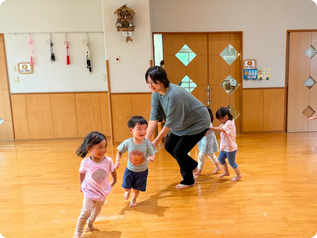 木目調の保育室で、保育士の後ろに並んで一列になり、体を動かして室内遊びを楽しむ子どもたちの様子