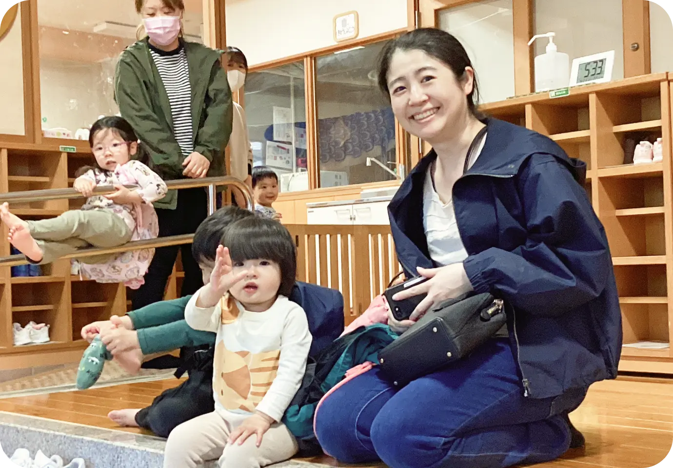 保育園の玄関・昇降口で、靴箱を背景に子どもたちと笑顔で座る保護者の様子