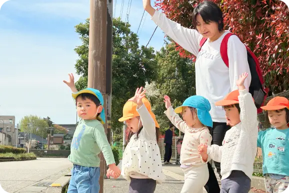 青空の下、保育士に手を引かれ、帽子をかぶって元気に外歩き(お散歩)をする子どもたちの様子