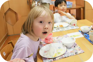 保育園の給食の時間に、スプーンを持ちながらカメラに向かって表情を浮かべる子どもの写真
