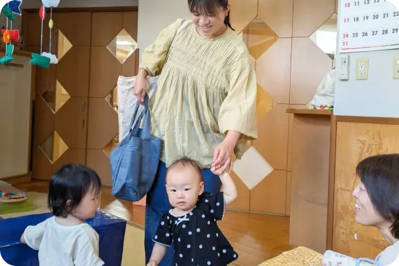 保育園の室内で、保護者に手を引かれて歩く乳児と、それを見守る保育士の様子