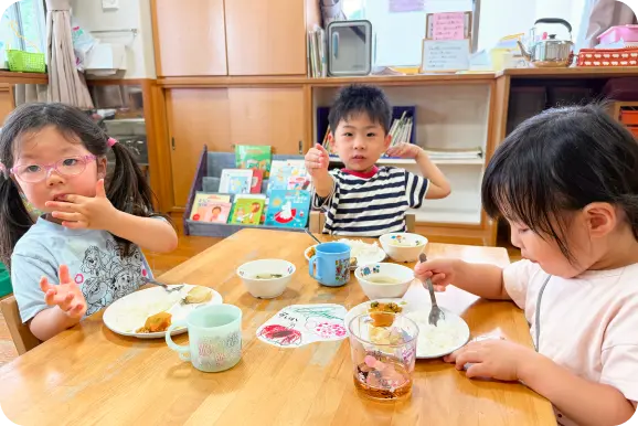 木目調の保育室で、テーブルを囲んで給食(食事)を食べる3人の子どもたちの様子