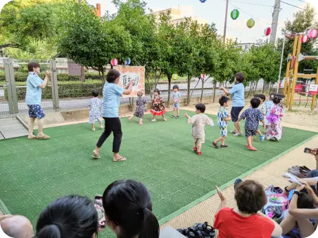 提灯が飾られた屋外の芝生広場で、浴衣や甚平を着て保育士と一緒に盆踊りをする子どもたちの様子