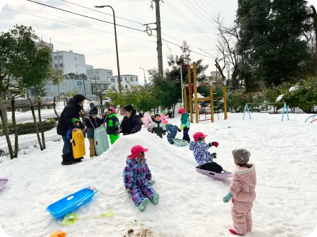 雪が積もった屋外の広場で、スキーウェアを着てそり遊びや雪山での活動を楽しむ子どもたちの様子