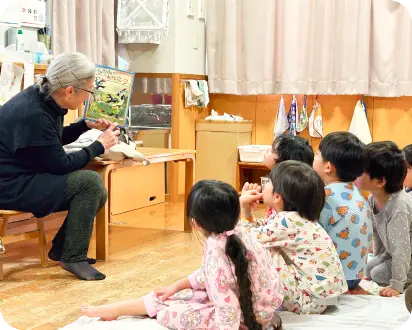 保育室で床に座る子どもたちに向けて、椅子に座った年配の女性が絵本を読み聞かせている様子