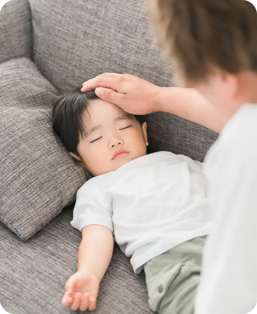 ソファの上で横になって目を閉じている子どもの額に、大人が手を当てている様子