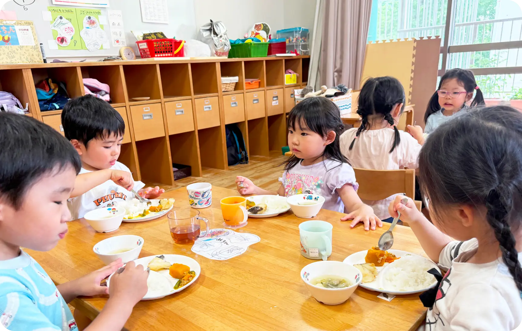 保育室の木製テーブルを囲み、スプーンを使って給食を食べる子どもたちの様子