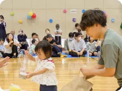 体育館で行われている行事で、大人の男性からプレゼントの袋を受け取る小さな子どもの様子