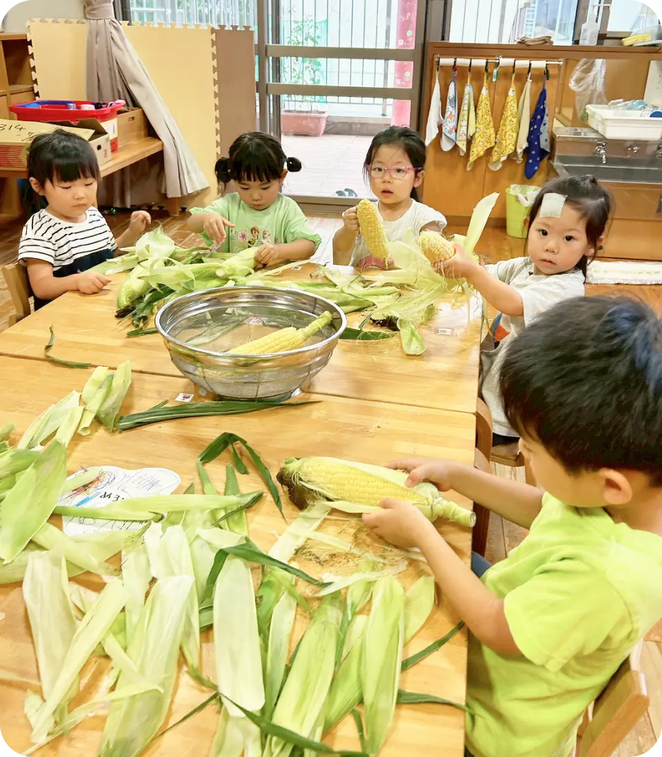 保育室のテーブルを囲み、並んでとうもろこしの皮むきを行っている子どもたちの様子