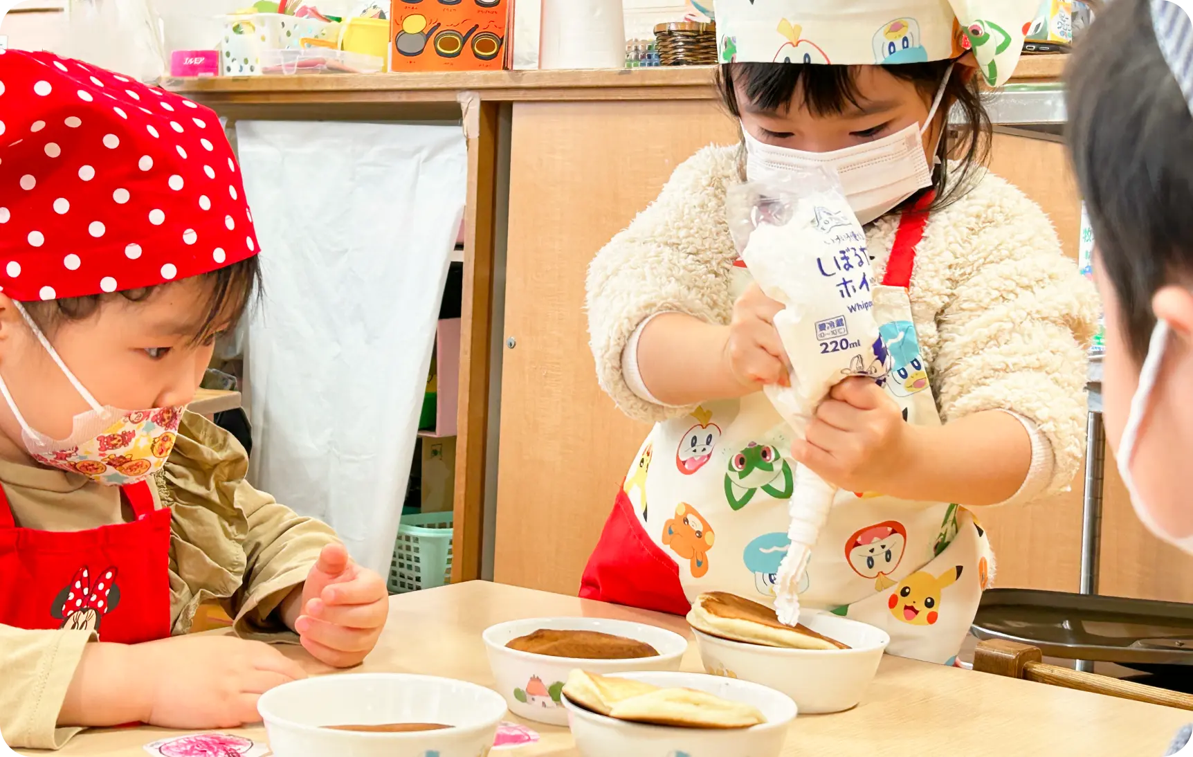 エプロンとマスクを着用し、パンケーキの上に自分でホイップクリームを絞り出す子どもの様子