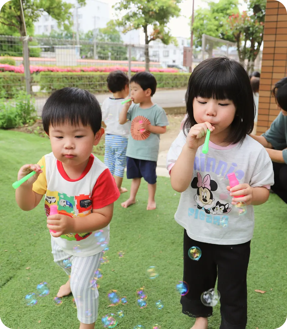 保育園の園庭にある人工芝の上で、スティック状の道具を使ってシャボン玉を吹いて遊ぶ子どもたちの様子