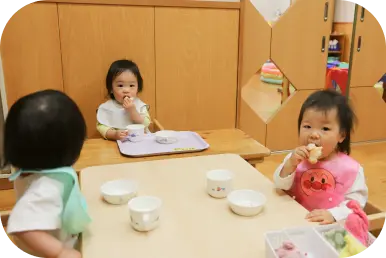 乳幼児用のローテーブルと椅子に座り、エプロンをつけておやつや離乳食を食べる小さなお子様たちの様子