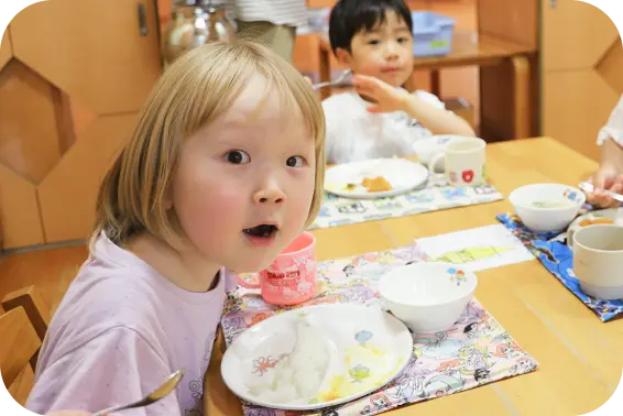 保育園の給食の時間に、スプーンを持ちながらカメラに向かって表情を浮かべる子どもの写真
