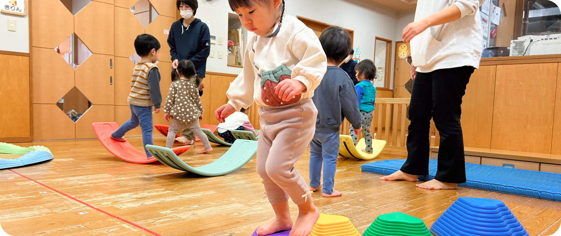 木目調の保育室の中で、カラフルなバランスボードや飛び石の遊具を使って室内遊びを楽しむ子どもたち