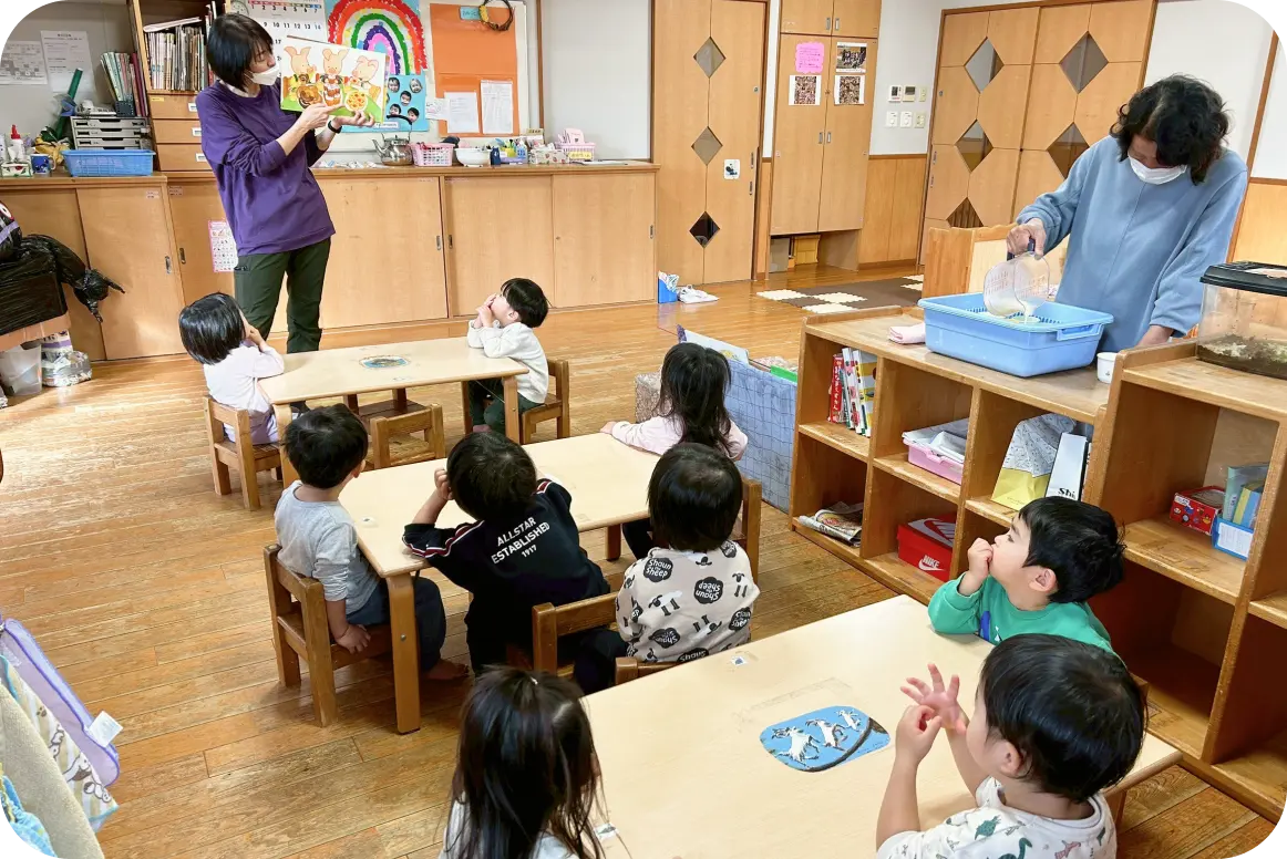 木目調の保育室で、テーブル席に座り保育士の絵本の読み聞かせを真剣に聞く子どもたちと、傍らで作業をする別の保育士の様子