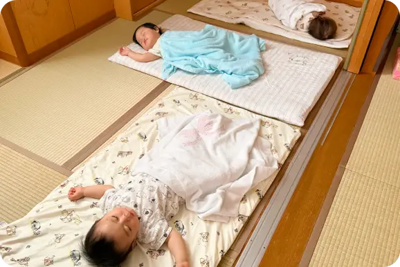 和室の保育室でお昼寝の時間に、布団に横になりタオルケットをかけられて眠る乳幼児たちの様子