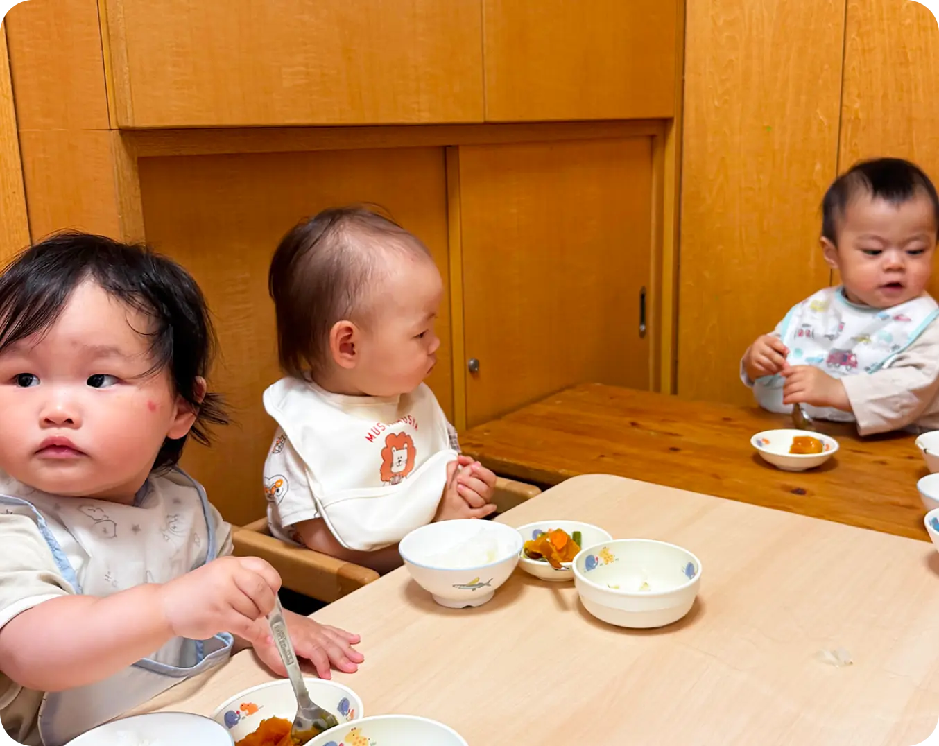 乳幼児用の高椅子に座り、それぞれのテーブルに並べられた給食を食べる小さな子どもたちの様子