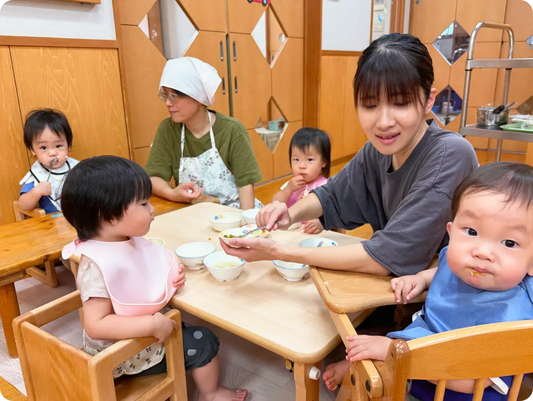 木目調の保育室で、エプロンをつけた保育士に見守られながら、テーブル席で座って給食を食べる子どもたちの様子