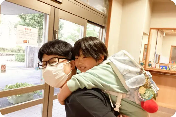 保育園の玄関・昇降口付近で、メガネとマスク姿の保護者に笑顔でおんぶされる子どもの様子