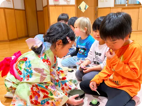 和室の保育室で、着物を着た子どもがお茶を点てて振る舞い、受け取った子どもたちが和菓子を食べるお茶会体験の様子