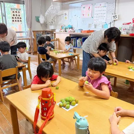 保育室のテーブルに座り、保育士と一緒に青梅のヘタ取り作業（梅仕事）をしている子どもたちの様子