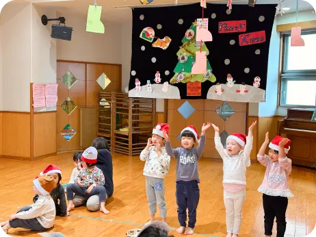 クリスマスツリーの壁面装飾がある室内で、サンタクロースの帽子をかぶって並び、歌やダンスを披露する子どもたちの様子