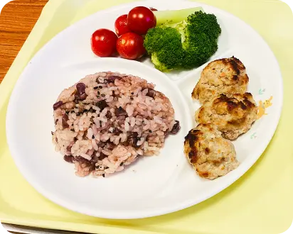 白い仕切り皿に盛り付けられた、お赤飯、ハンバーグ(つくね)、ミニトマトとブロッコリーの給食メニュー