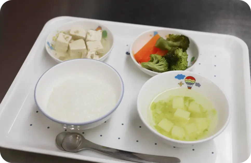 白いトレイの上に並べられた、お粥やスープ、豆腐、温野菜が入ったアレルギー対応食の画像
