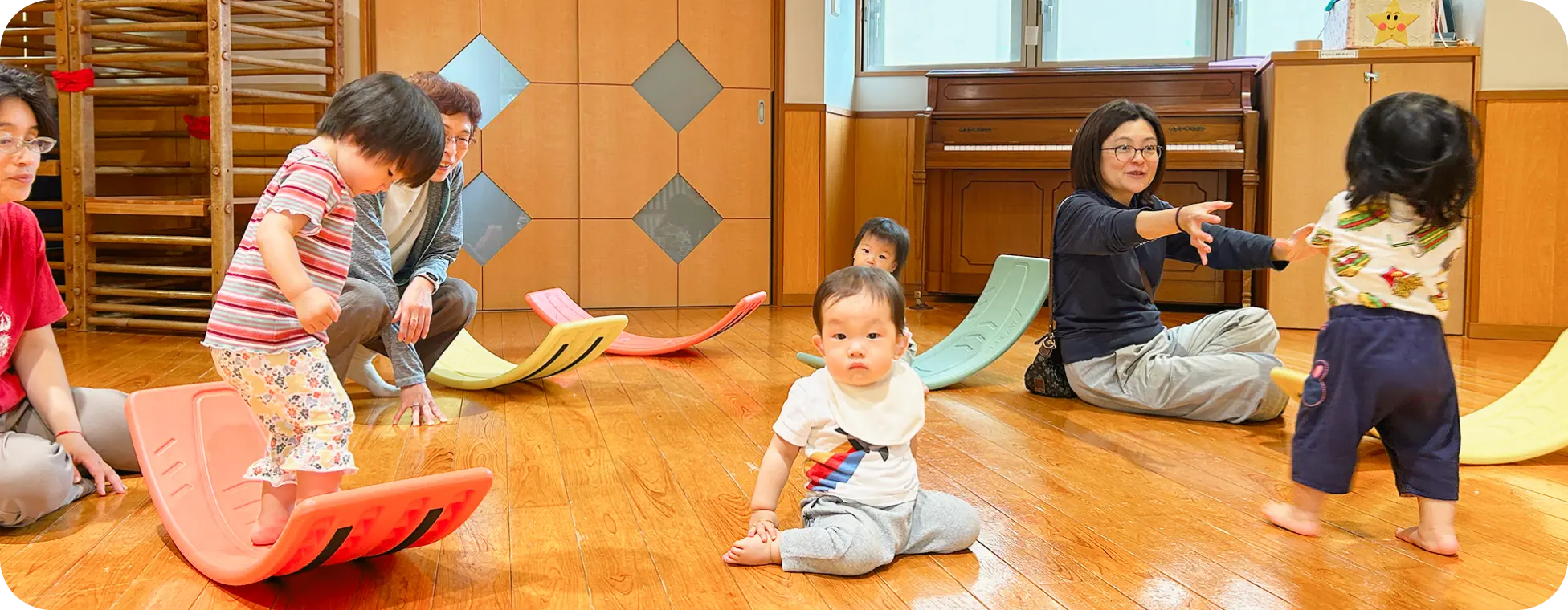 保育園の遊戯室で、カラフルなバランスボードに乗って遊ぶ子どもたちと、それを見守りサポートする保育士の様子