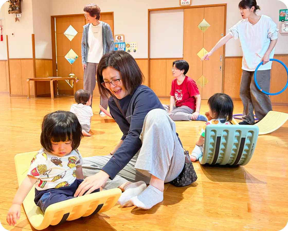 保育園の遊戯室で、バランスボードに乗る子どもの近くに座りサポートする保育士と、周囲で見守る大人たちの様子