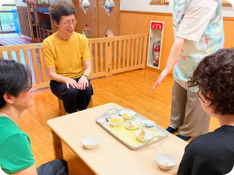 保育室の小さなテーブルを囲み、エプロン姿の職員を交えて離乳食の試食や会話をする大人たちの様子