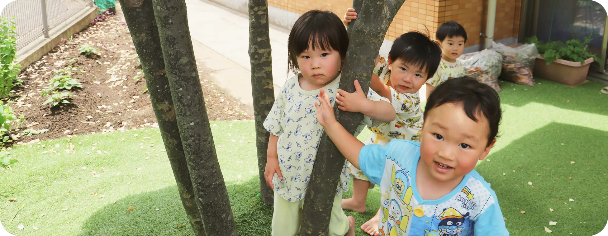 保育園の園庭にある木の周りに集まり、カメラに向かって微笑む子どもたちの様子