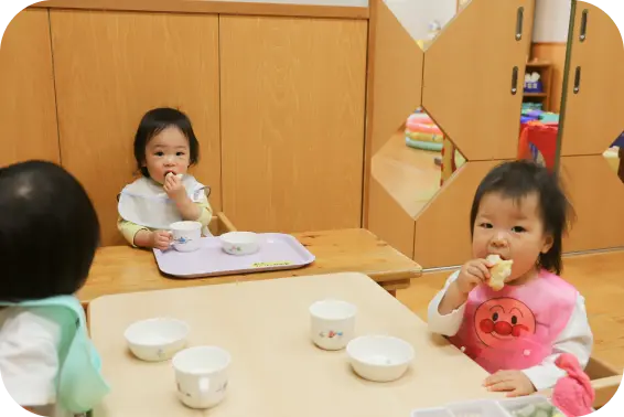乳幼児用のローテーブルと椅子に座り、エプロンをつけておやつや離乳食を食べる小さなお子様たちの様子