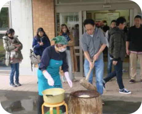 園のイベントで、子どもたちや保護者が見守る中、大人たちが餅つき（もちつき）体験を行っている様子