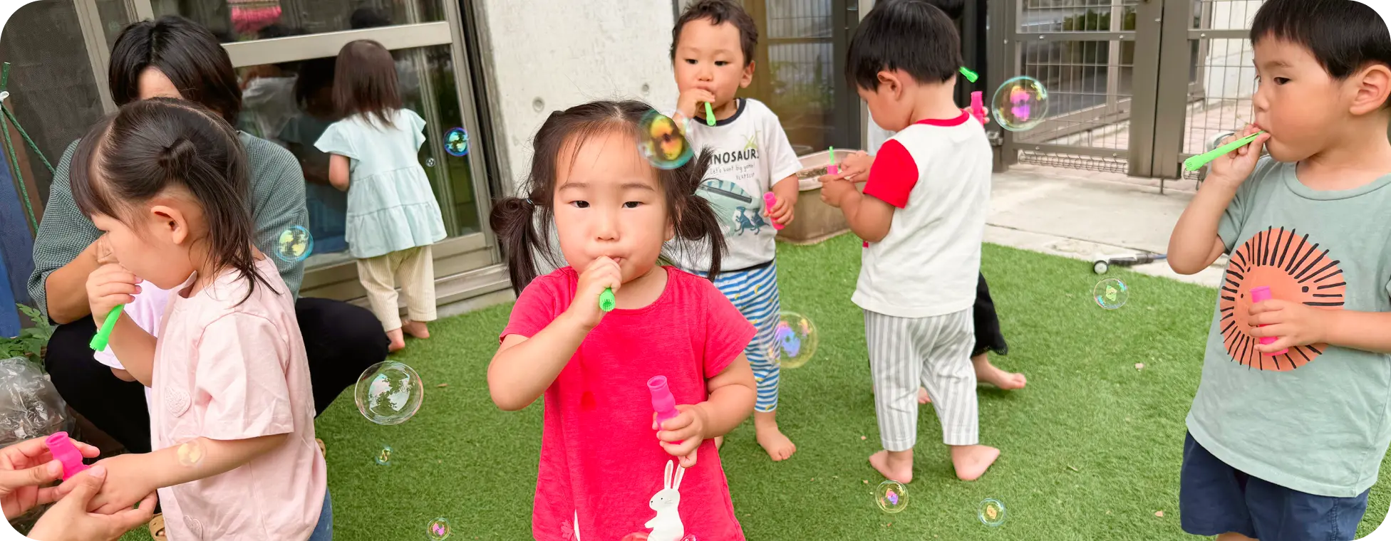 保育園の園庭にある人工芝の上で、集まってシャボン玉を吹いて遊ぶ子どもたちの様子
